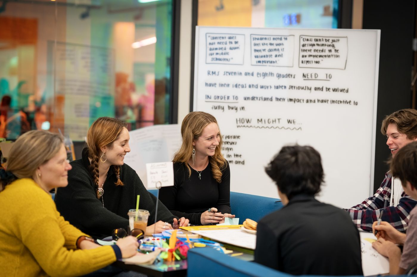 Students and educators around a table, working under a whiteboard that reads “How might we…”
